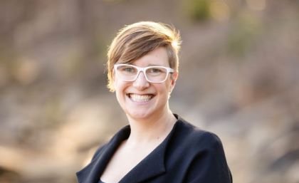 A woman with short hair in a black jacket and white glasses smiles broadly.